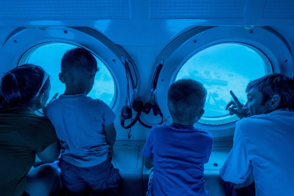 Vier Kinder knien an zwei runden U-Boot-Bullaugen und schauen in blaues Wasser