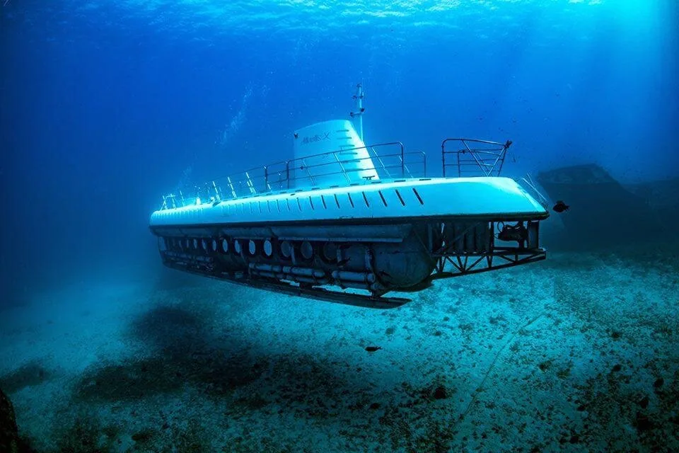 A white submarine cruising over a sandy, rocky sea floor in blue water