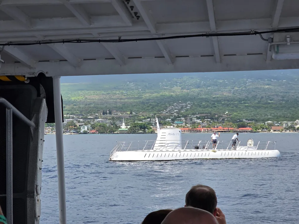 Gästefoto des weißen U-Boots an der Oberfläche, dahinter die Stadt Kailua-Kona und grüne Hügel